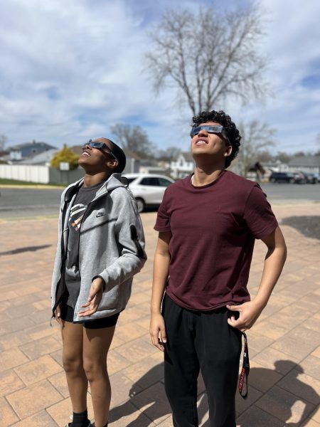 Junior Mia Montanez and senior Maanav Amin test out solar eclipse glasses in front of JFKMHS.